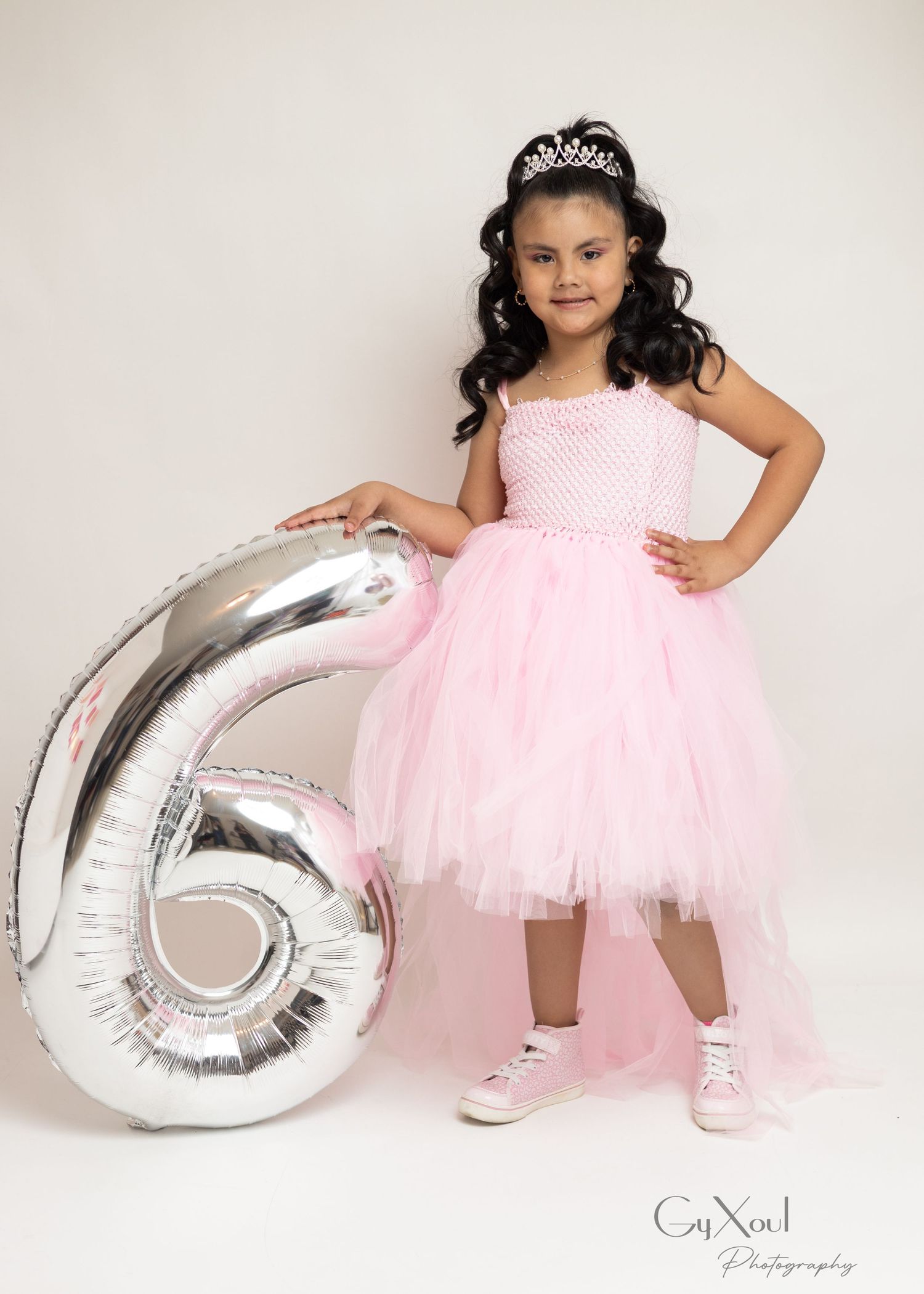 A young girl in a pink tulle dress and tiara poses beside a silver number 6 balloon for her birthday photo.
