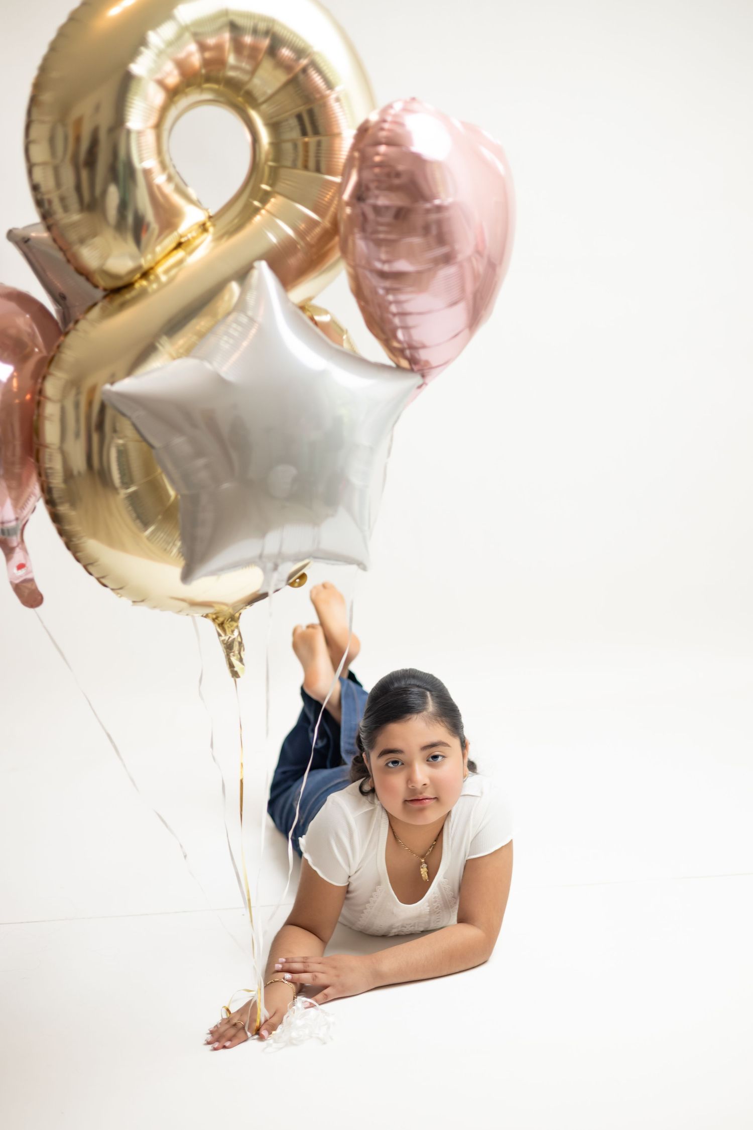 Young girl lying on white floor holding gold number 8, star, and heart-shaped foil balloons for birthday celebration.