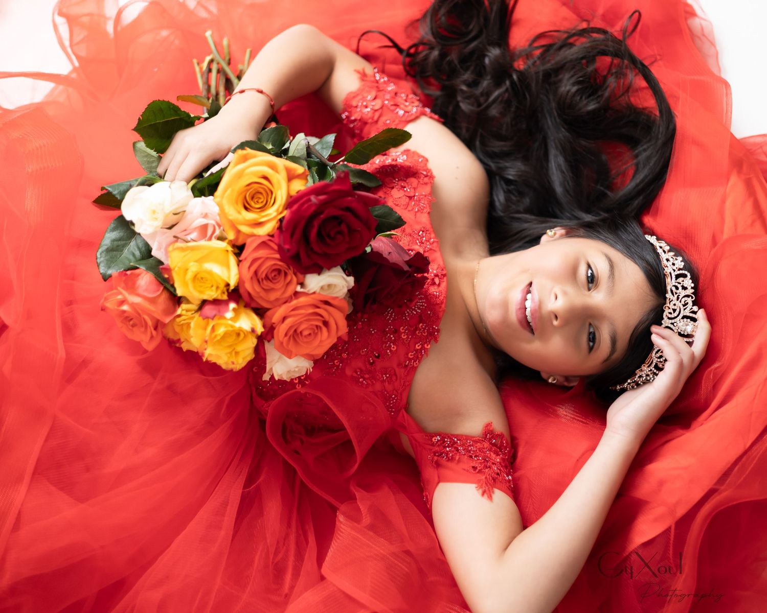 Young woman in red gown holding colorful roses bouquet, wearing tiara, lying down for quinceañera portrait photo.