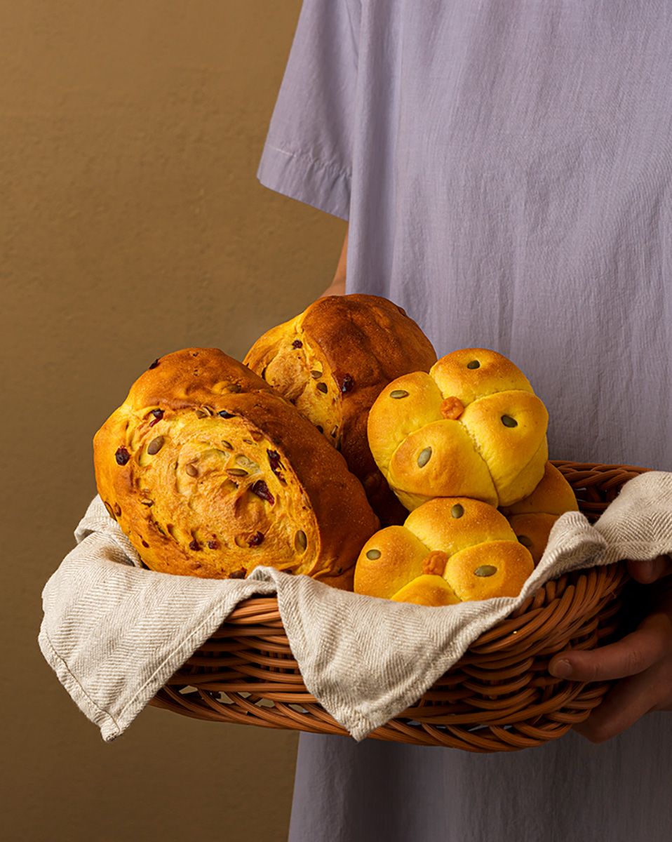 Person holding a wicker basket lined with linen cloth filled with golden bread rolls and rustic raisin loaves.
