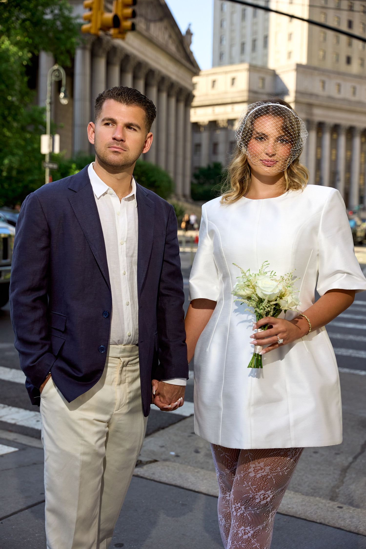 Newlywed couple holding hands on a city street, bride in white mini dress with veil and bouquet, groom in navy blazer.
