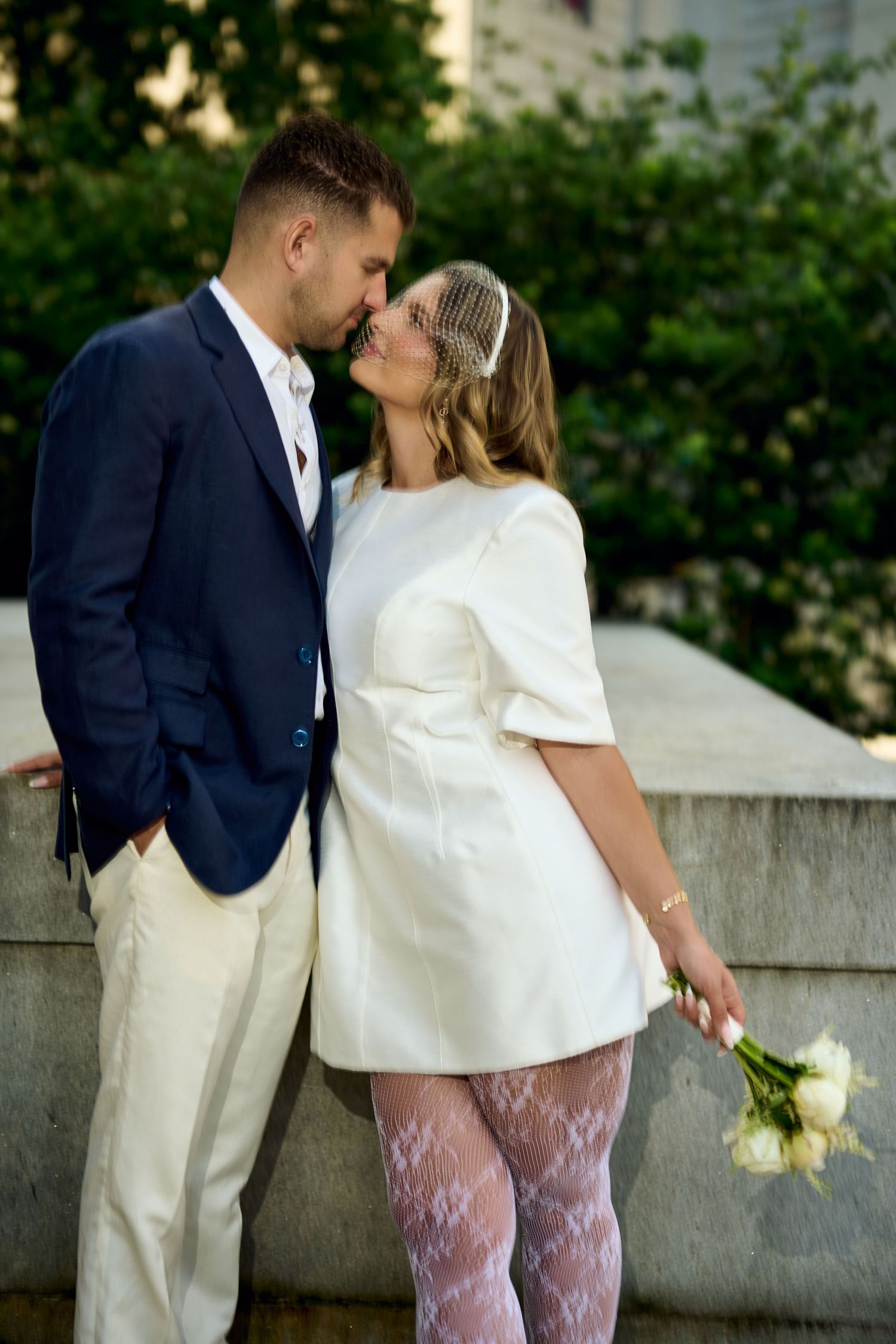 Couple in wedding attire kissing outdoors, bride in white mini dress with bouquet, groom in navy blazer.