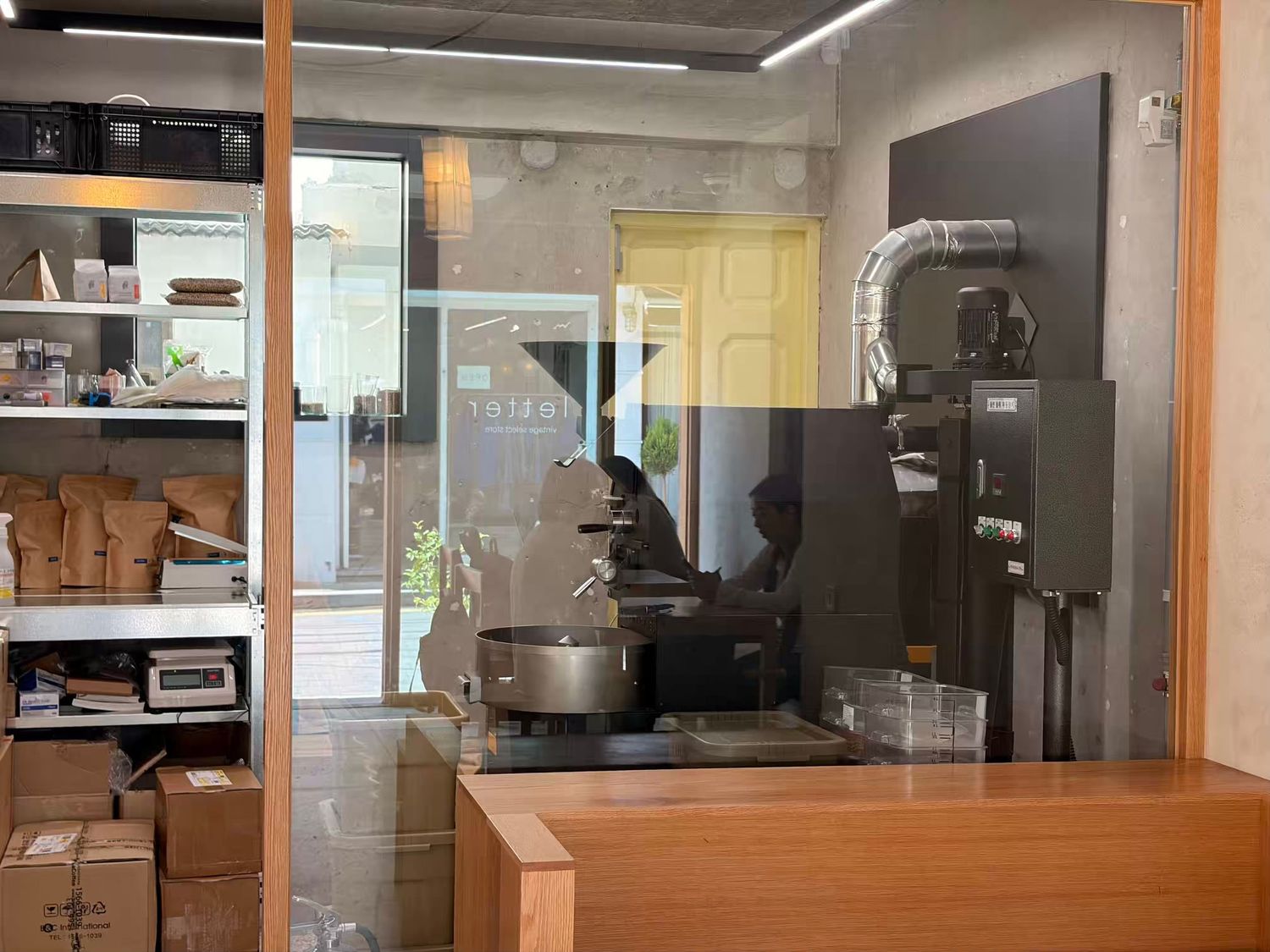 Interior view of a coffee roastery shop with a professional roasting machine behind glass partition and storage shelves.