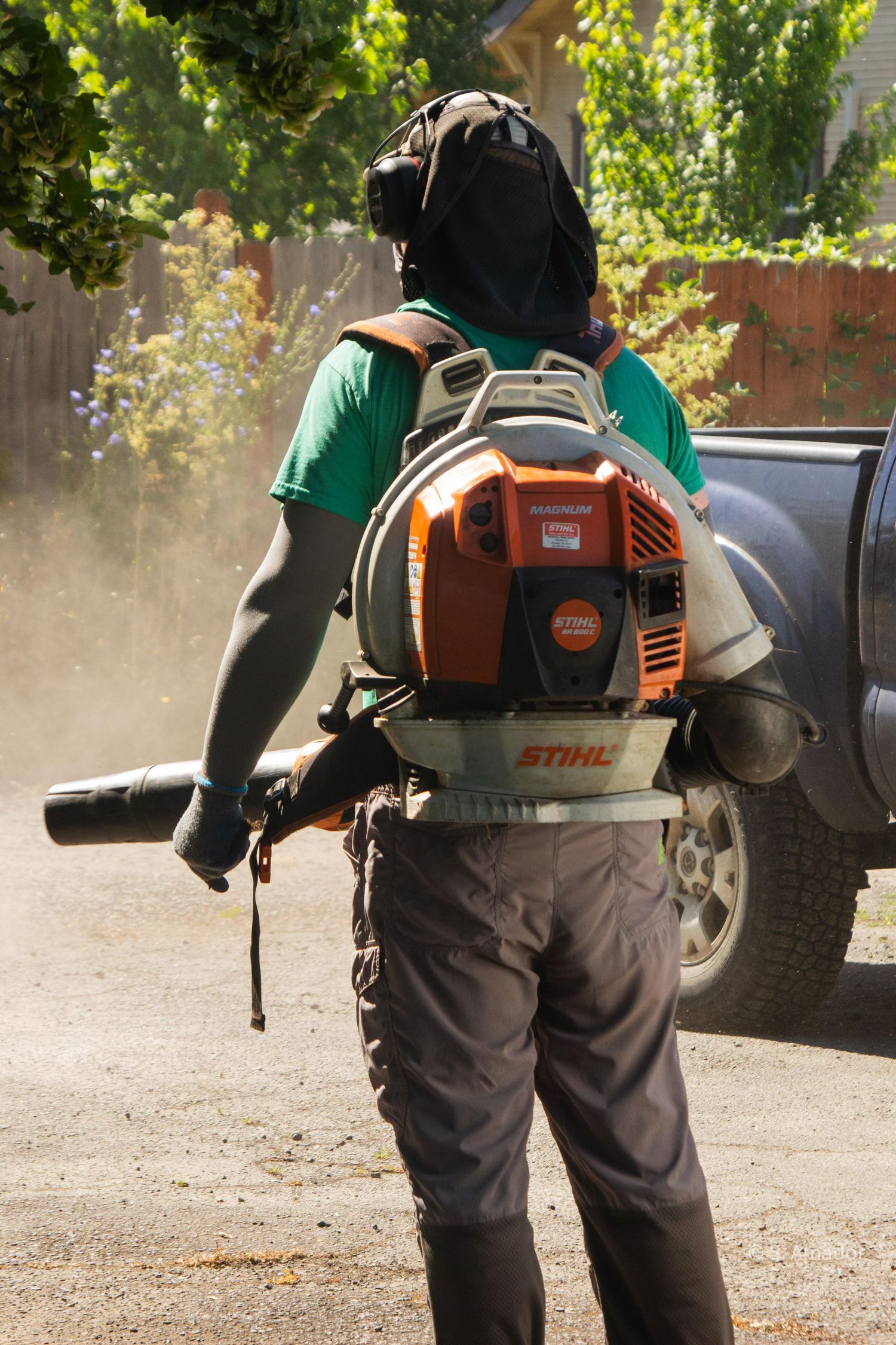 A worker wearing a backpack leaf blower and protective gear blows dust in a residential driveway.