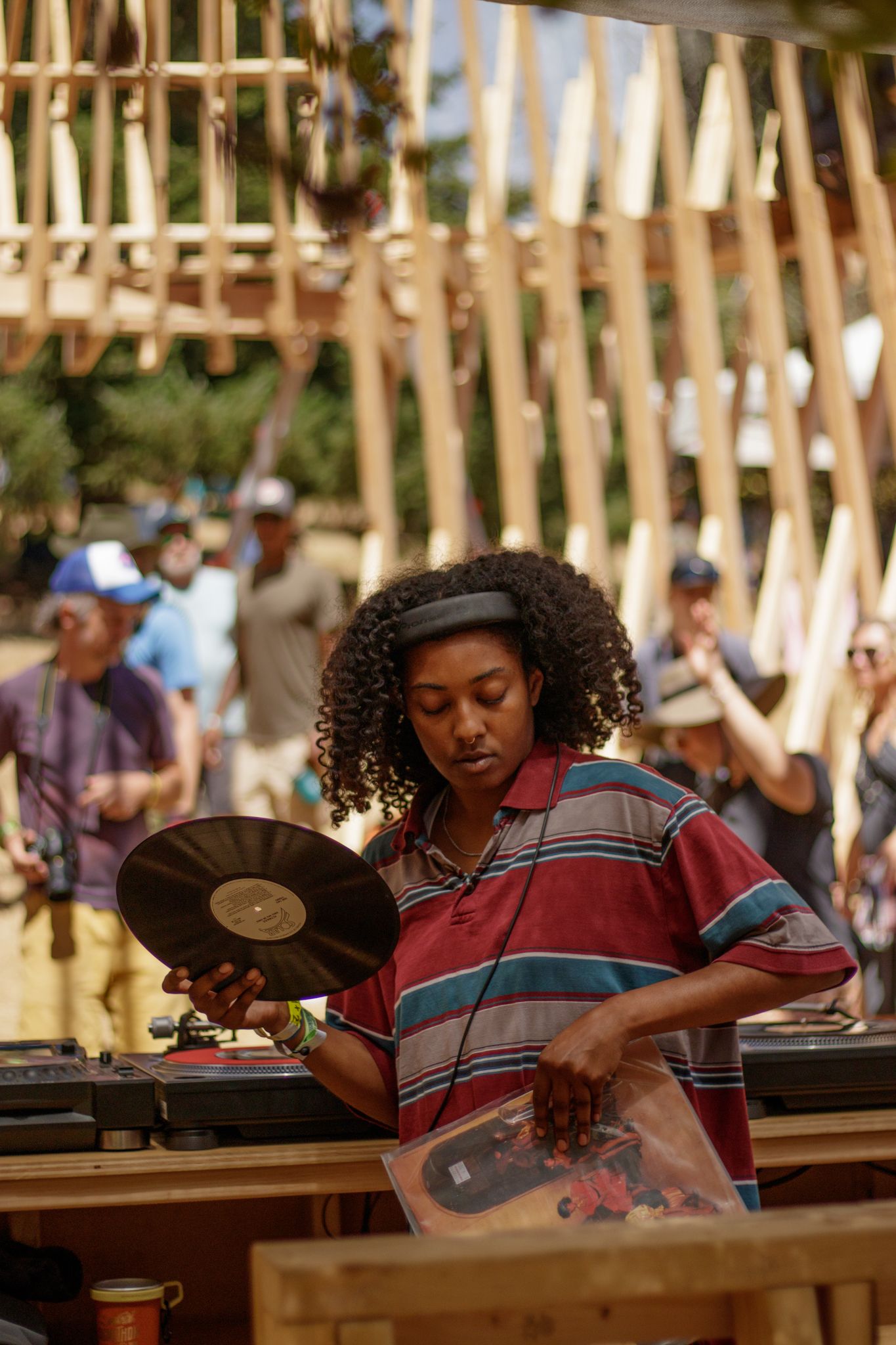 A woman with curly hair holds a vinyl record at an outdoor DJ setup during a sunny festival event.