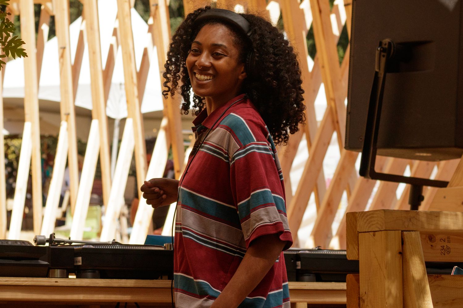Smiling young woman with curly hair wearing a colorful striped shirt, standing outdoors near wooden decor.