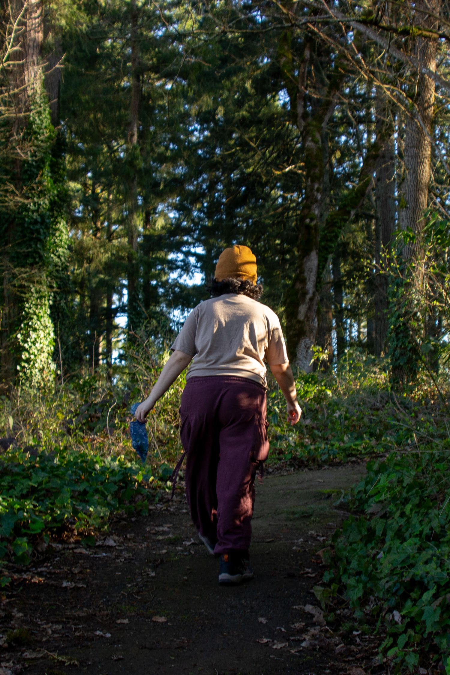 A person wearing a yellow beanie and maroon pants walks along a shaded forest path surrounded by tall trees.