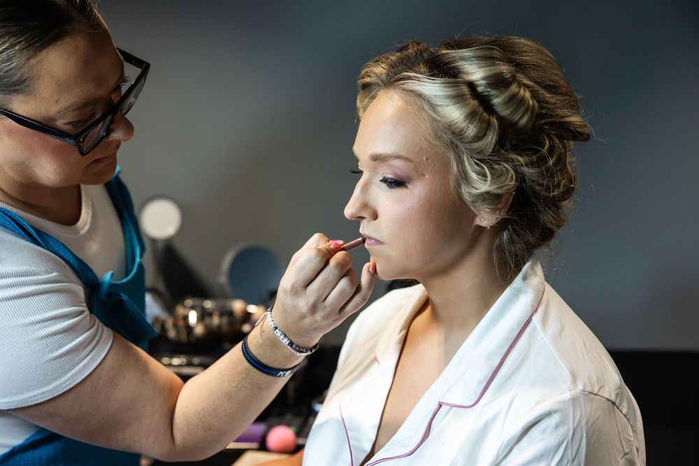 Makeup artist applying lipstick to blonde bride wearing white robe during wedding preparation.