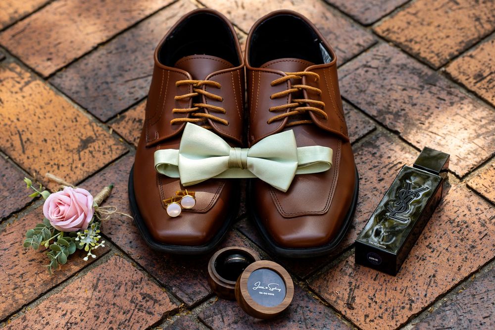 Brown leather dress shoes with gold lace, mint bow tie, pink rose boutonniere, cufflinks, and cologne on brick.