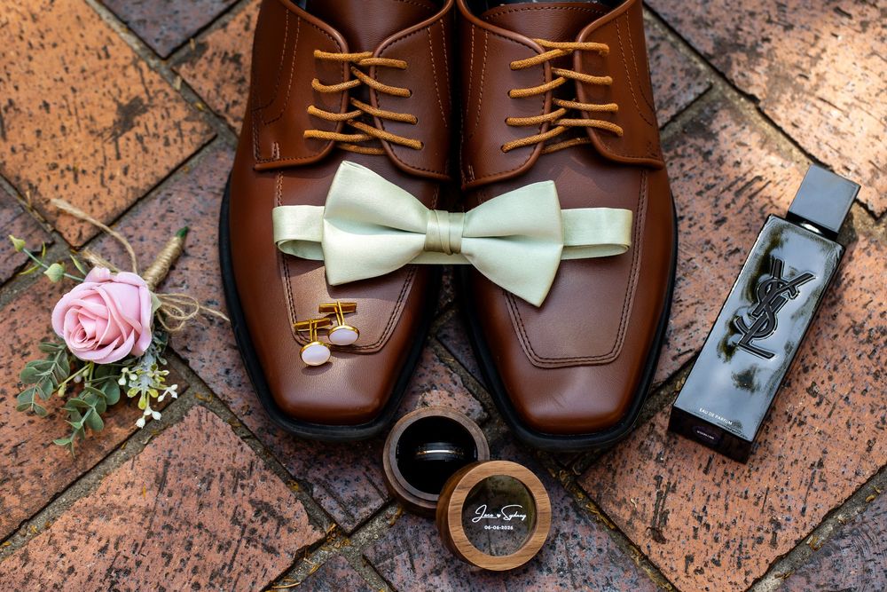 Brown leather dress shoes with mint bow tie, gold cufflinks, pink boutonniere, and cologne on brick surface.