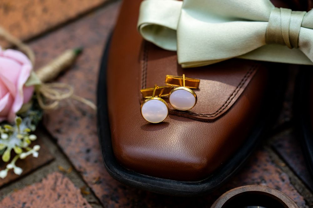 Gold circular cufflinks resting on brown leather dress shoe with mint bow tie and pink rose in background.