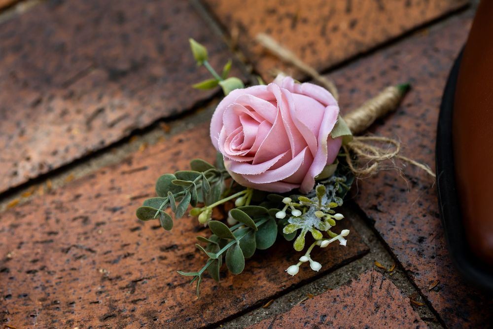 Pink rose boutonniere with greenery and twine on rustic brick surface for wedding day details.
