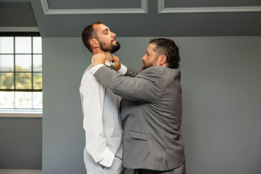Groomsman in gray suit helping groom in white suit adjust his bow tie before wedding ceremony.