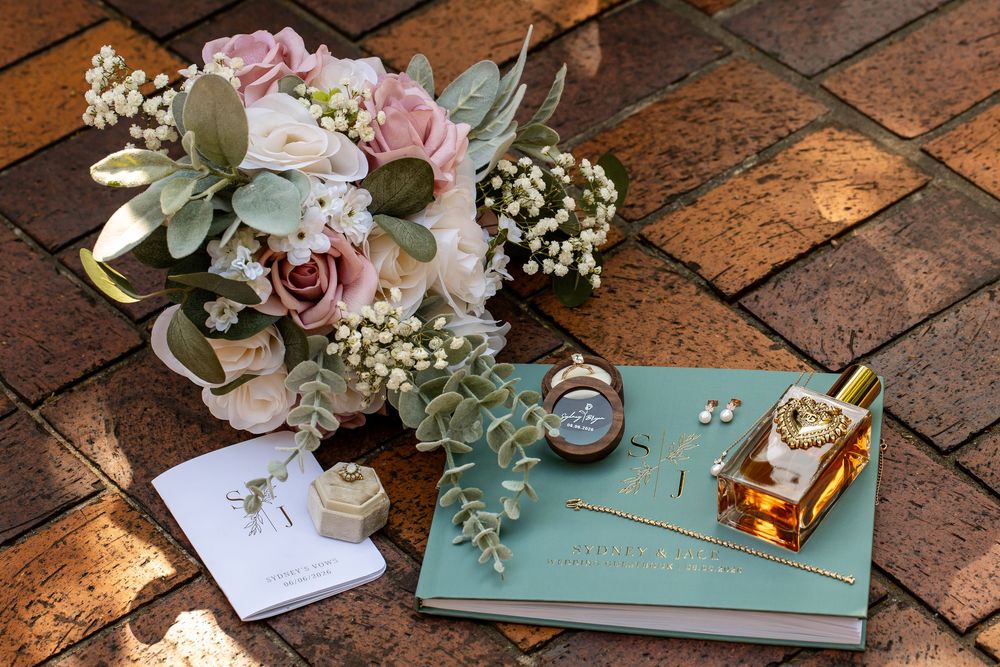 Wedding flat lay with pink rose bouquet, mint guestbook, perfume bottle, and accessories on brick surface.