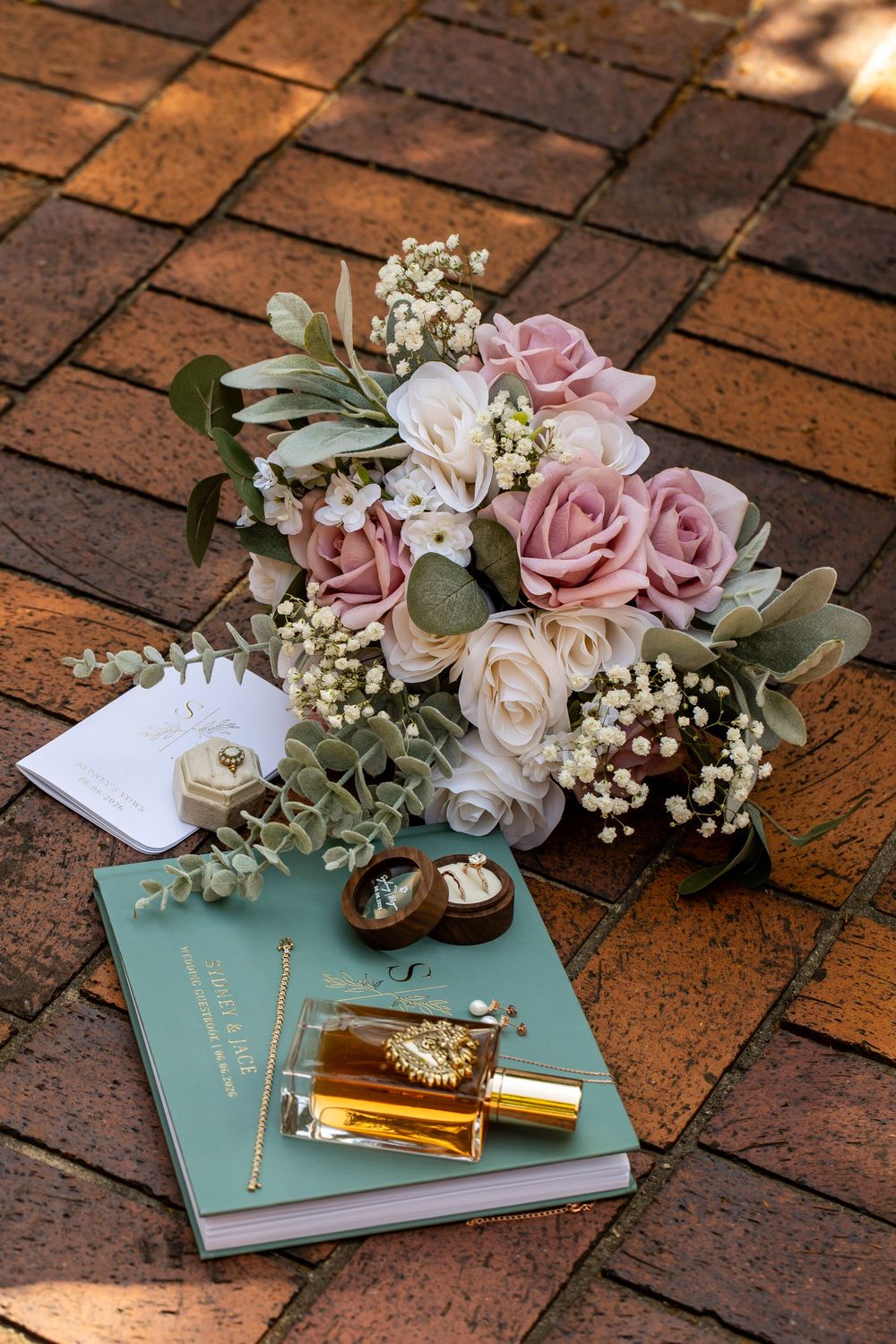 Elegant wedding flat lay featuring pink and white rose bouquet, mint guestbook, perfume, and ring box on brick.