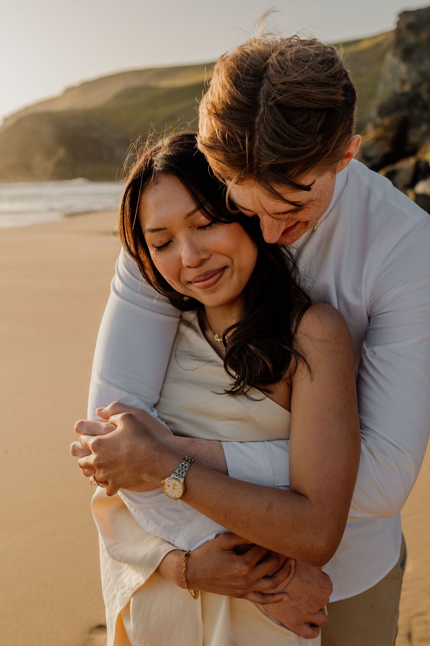Couple embracing on sandy beach at golden hour, with rocky cliffs in background, woman in white outfit.