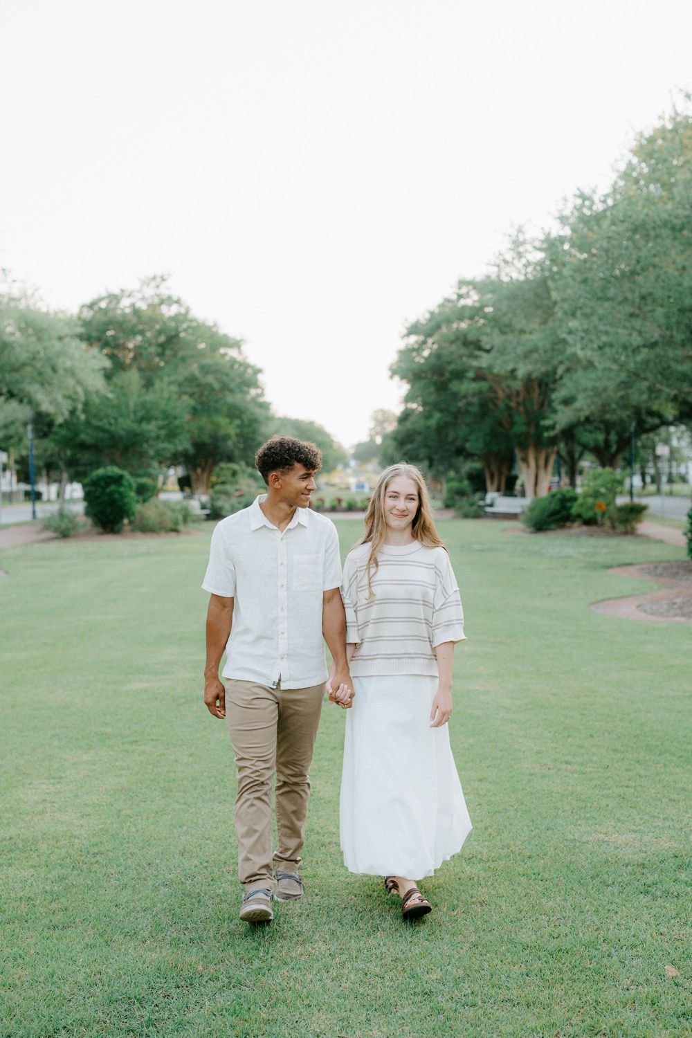 A young interracial couple holds hands while walking through a lush green park, dressed in casual neutral tones.