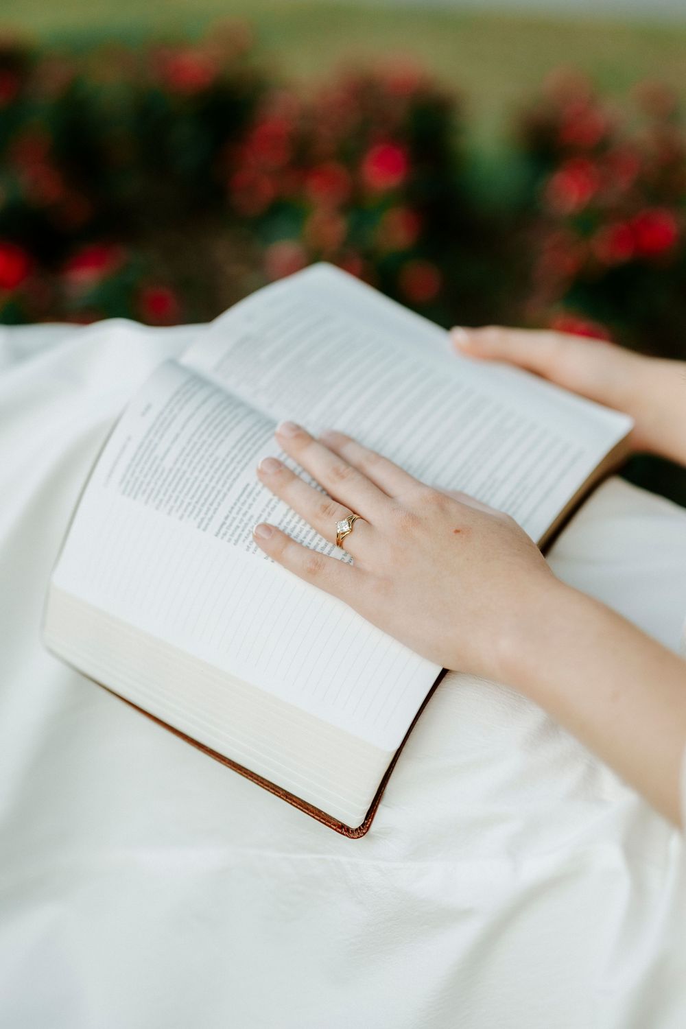 A woman wearing a gold ring rests her hand on an open book while sitting outdoors near red flowers.