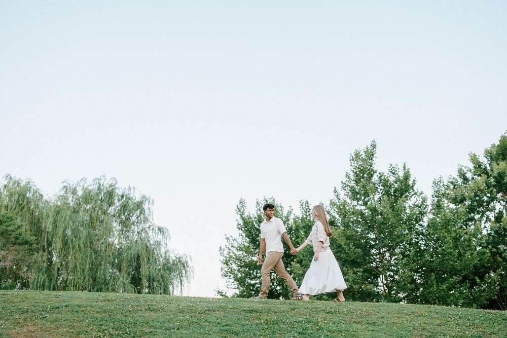 A couple holds hands while walking on a green grassy hill surrounded by lush trees on a clear day.