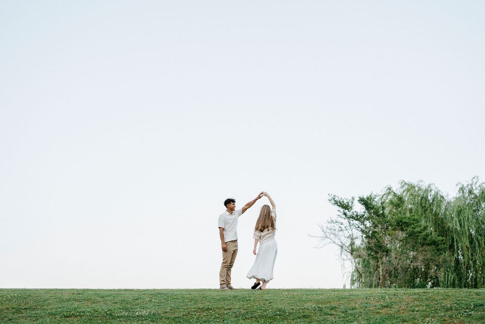 Couple dancing together on green grass field with trees and clear sky in the background.