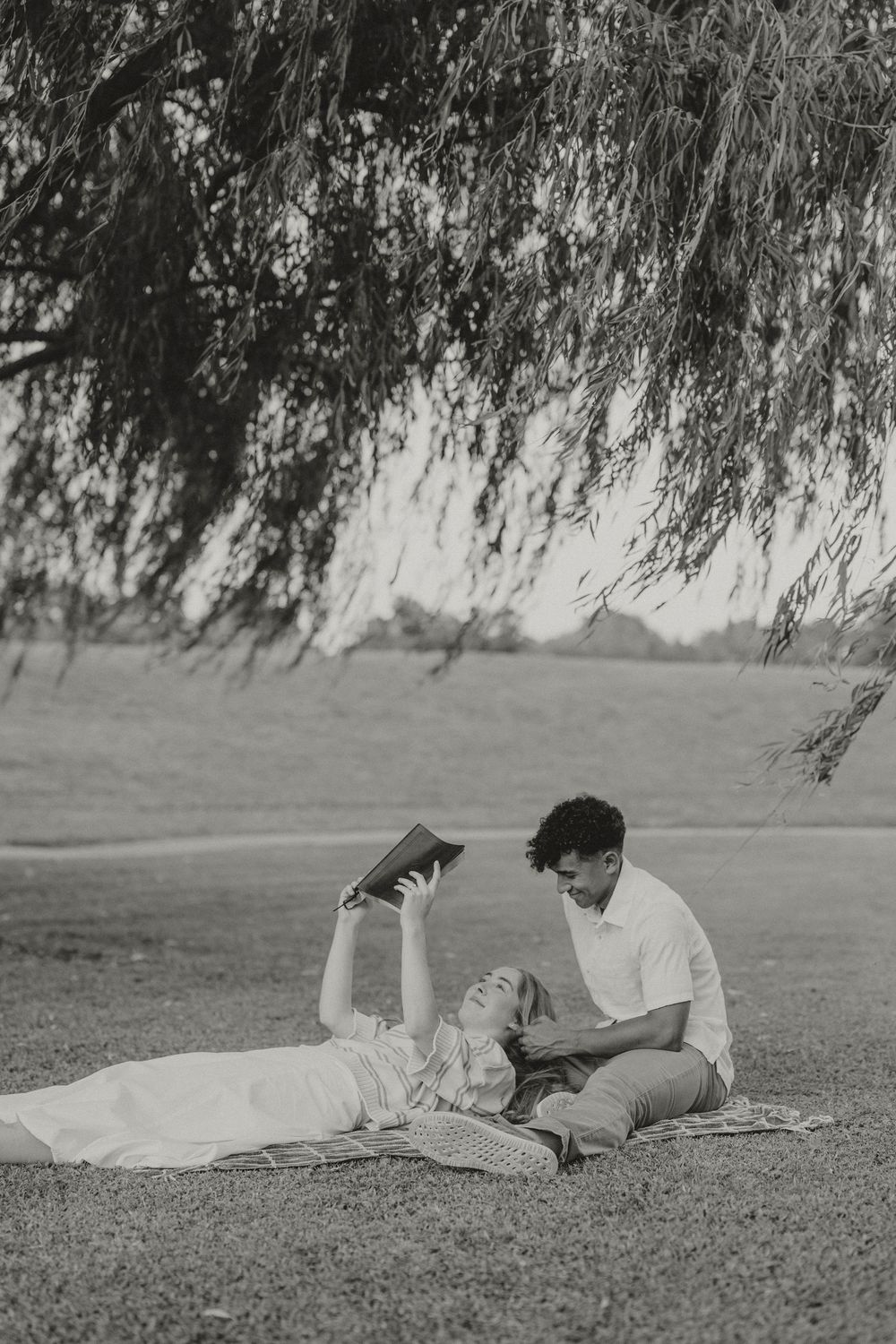 A couple relaxes under a willow tree in a black and white photo, reading books together on a blanket.