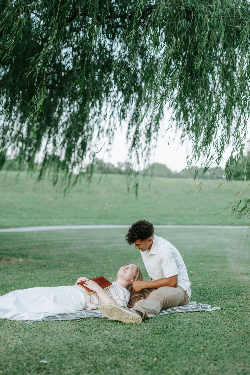 Couple relaxing under a willow tree in a green park, woman lying in man's lap holding a book.