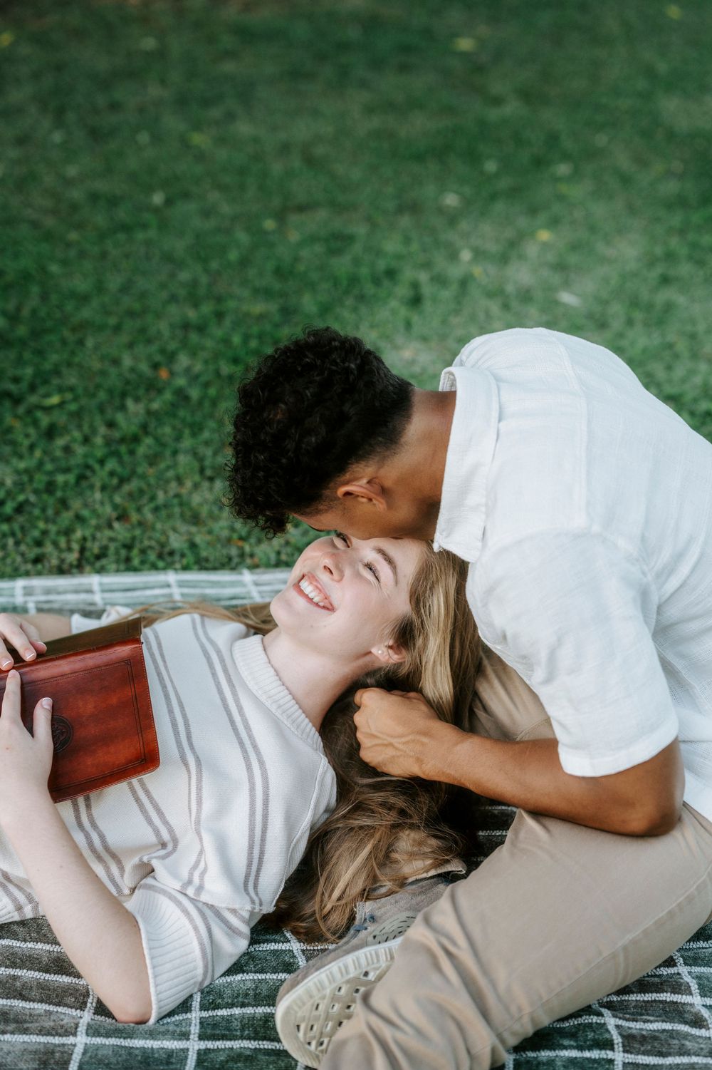 A joyful interracial couple relaxing on a blanket in a park, sharing a tender kiss while the woman laughs holding a book.