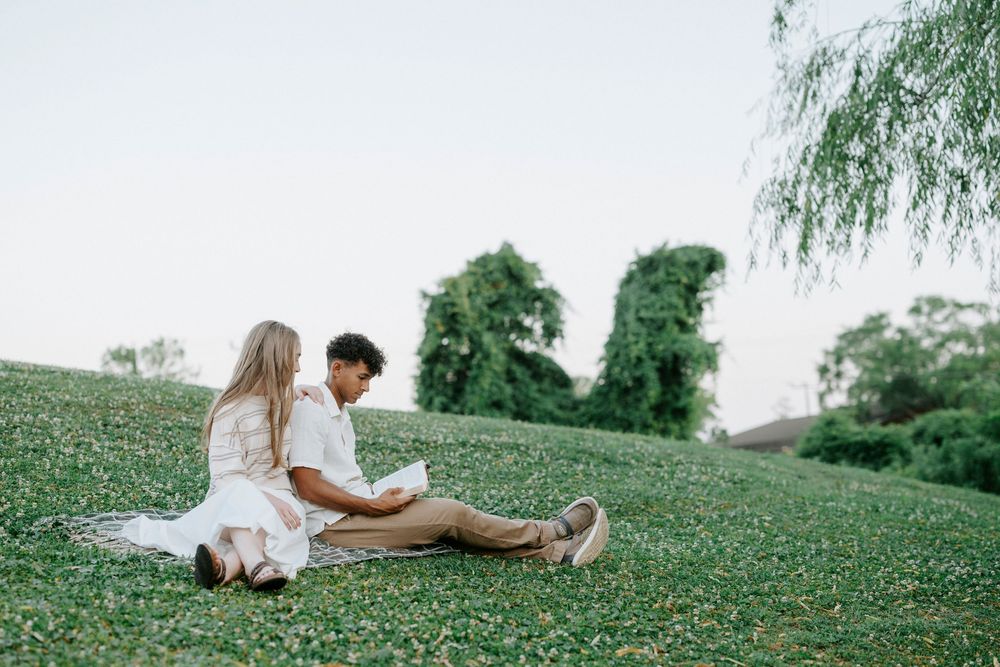 Couple sitting together on a lush green hillside, sharing a peaceful moment outdoors near trees.