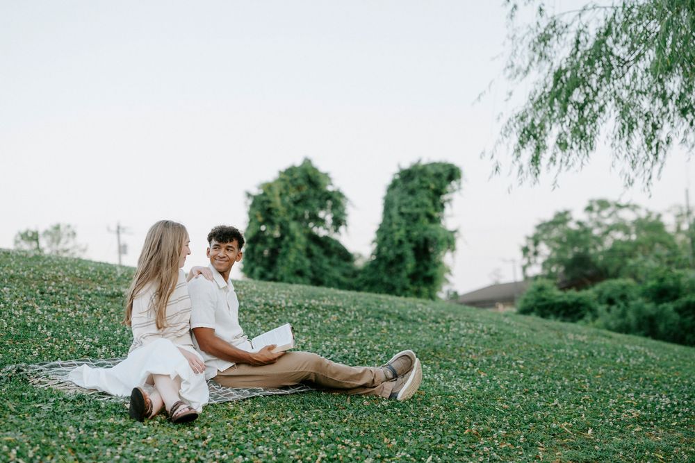 Couple sitting together on a green grassy hill outdoors, wearing white outfits on a sunny day.