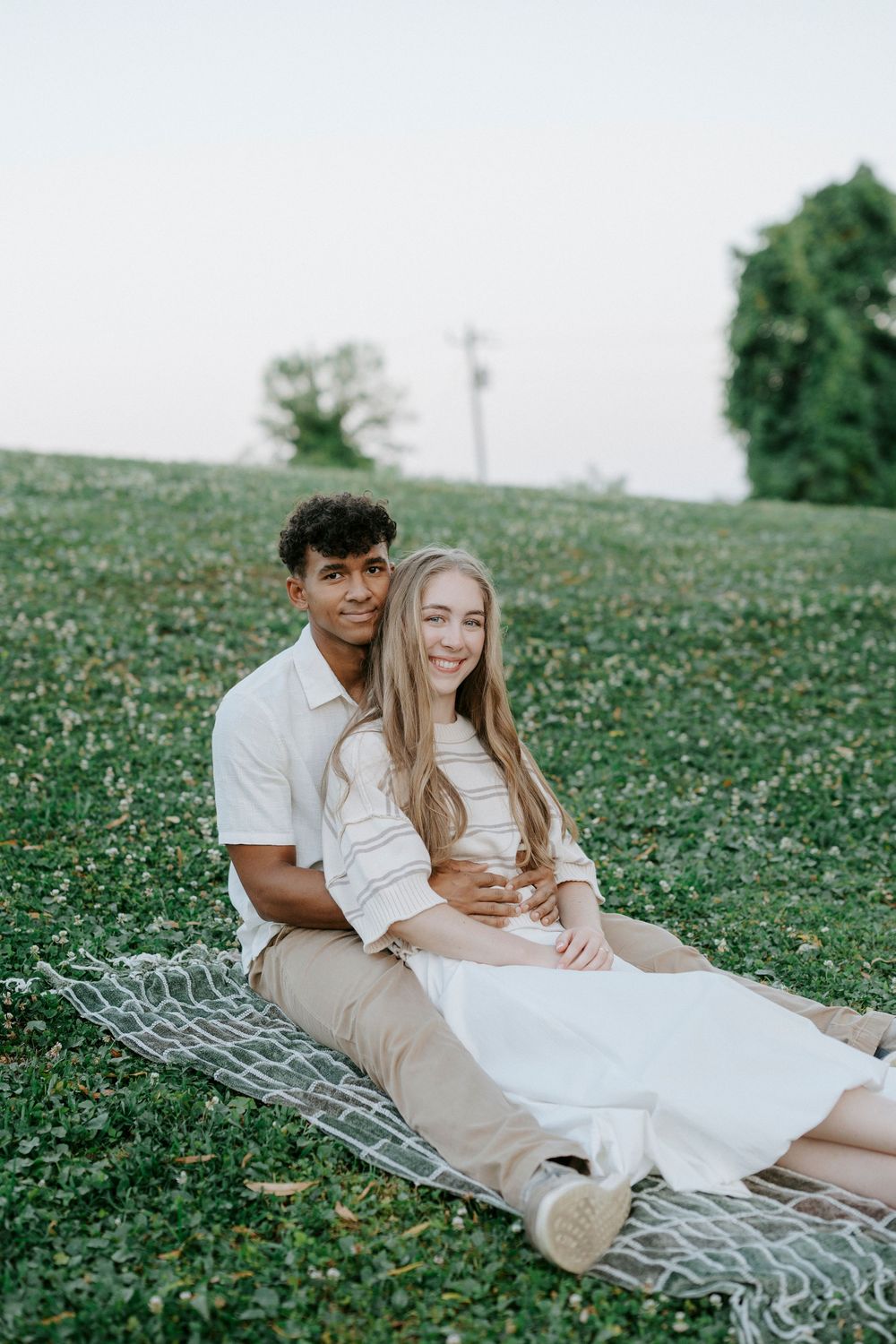 A young interracial couple sits together on a blanket in a green park, smiling warmly at the camera.