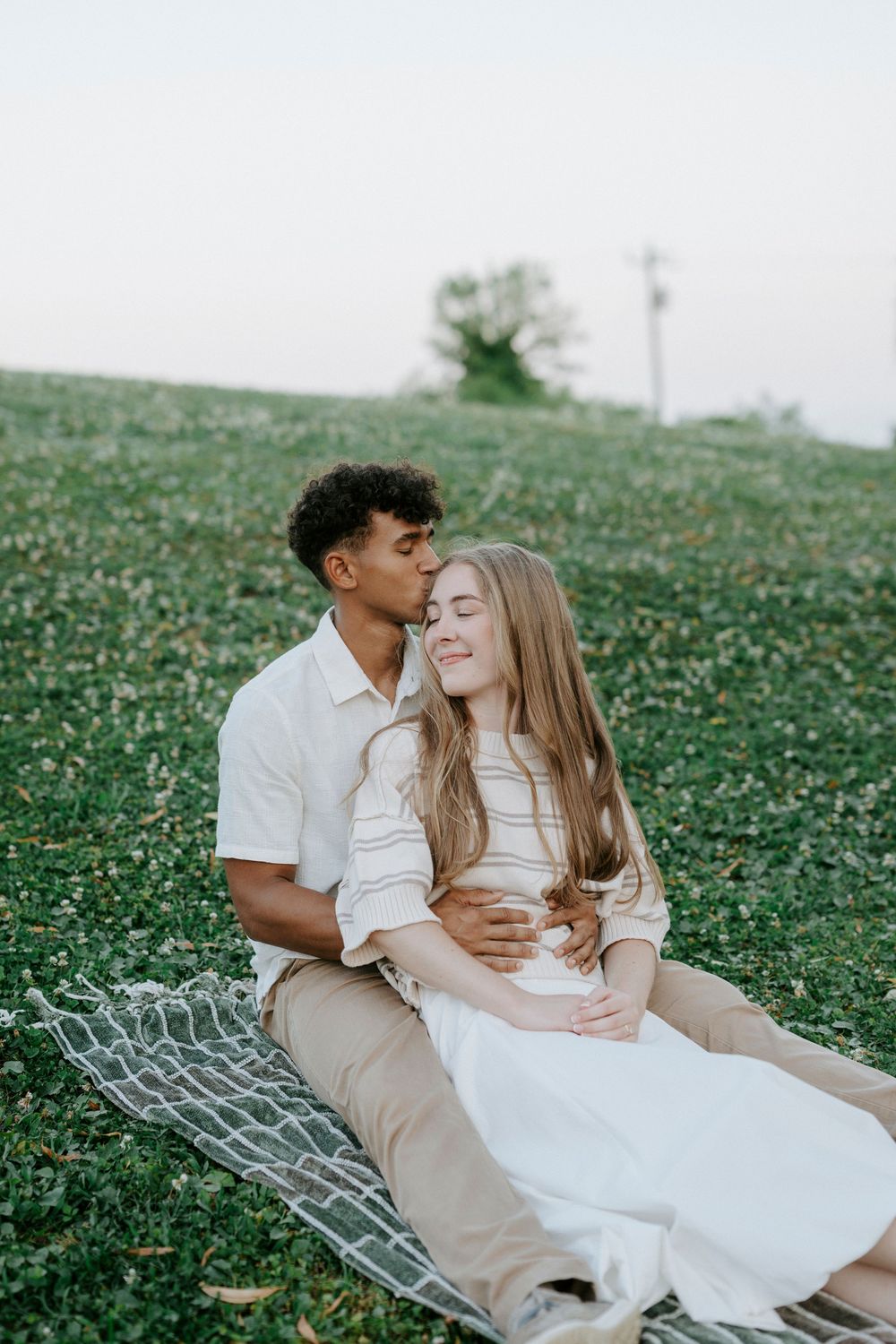 A young interracial couple sits on a blanket in a green field, sharing a tender kiss outdoors.