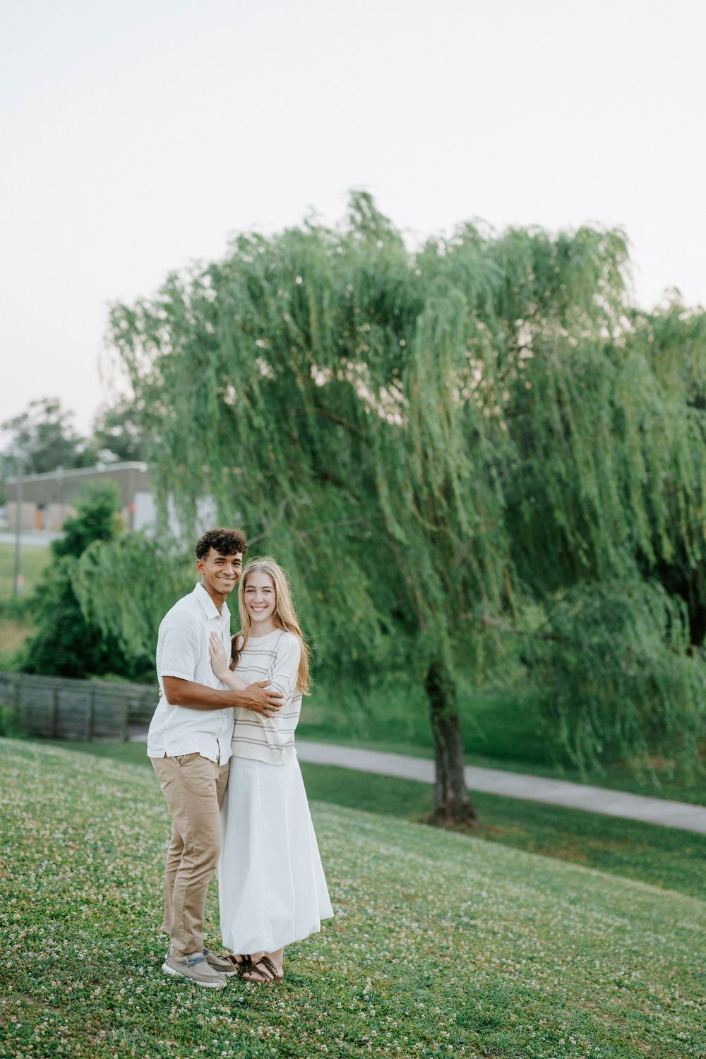 A smiling interracial couple embraces on a green hillside with a large weeping willow tree in the background.