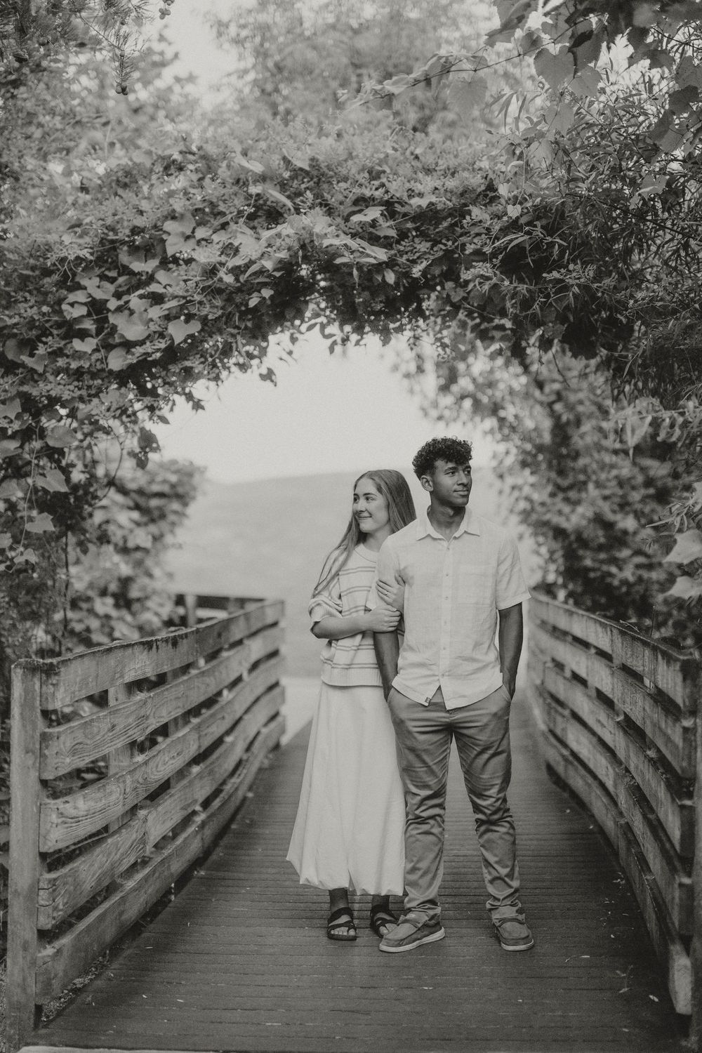 Black and white photo of a couple standing on a wooden bridge under a leafy arch, sharing a tender moment.