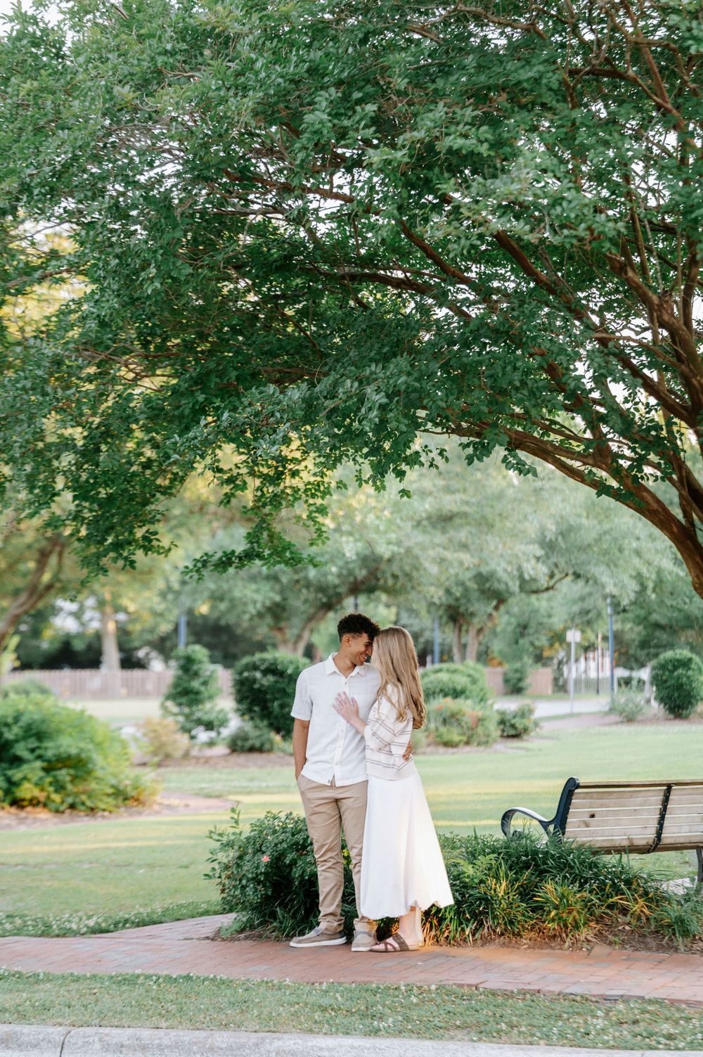 A romantic couple shares an intimate moment on a sunlit park path, surrounded by lush green trees and manicured gardens.