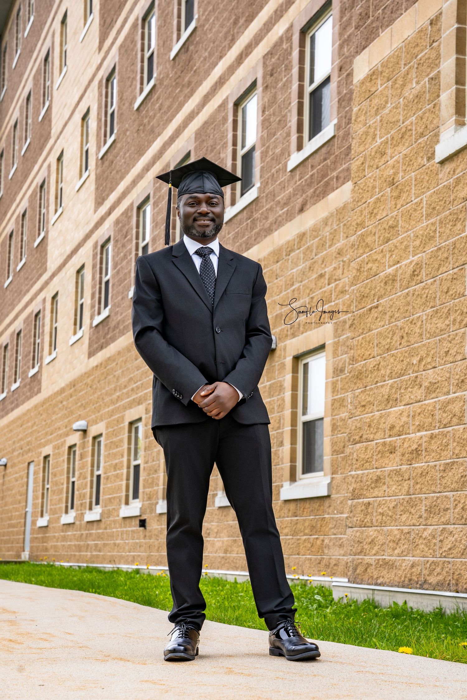 Smiling graduate in black cap and suit stands proudly outside a brick university building on graduation day.