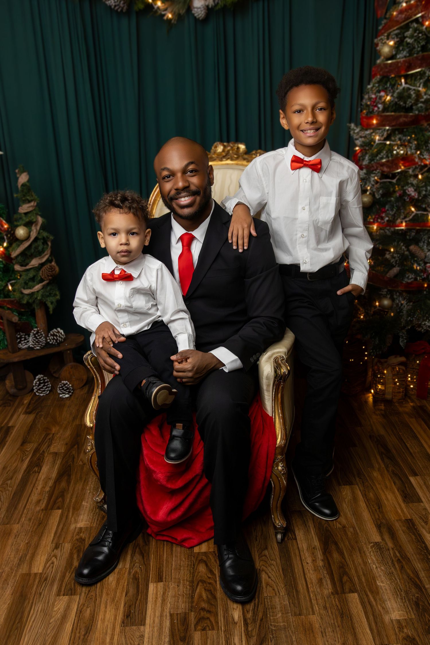 A man in a black tux and red tie sitting on a white ornate chair with his 2 sons, a toddler and a school aged child, all smiling
