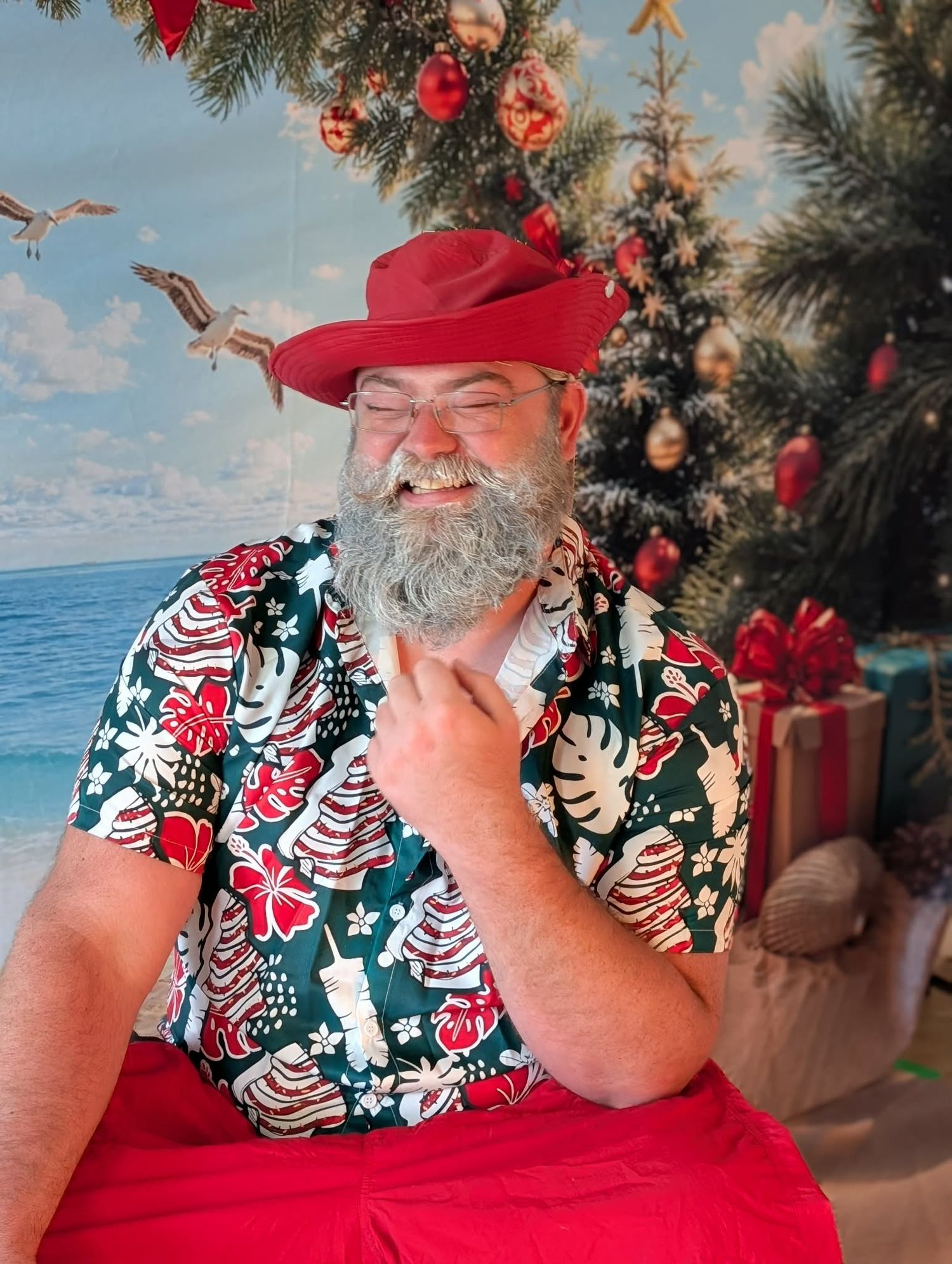Man with gray beard wearing red hat and Christmas Hawaiian shirt laughing near holiday tree and gifts.