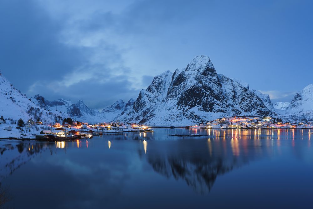 Snow-covered Norwegian fishing village glows at dusk, reflected in calm fjord waters with dramatic mountain backdrop.