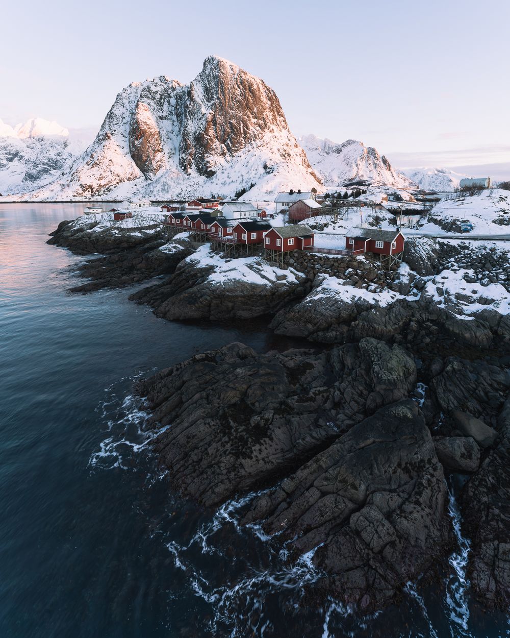 Aerial view of red Norwegian fishing cabins on snowy rocky shore with dramatic snow-capped mountain backdrop in Lofoten.