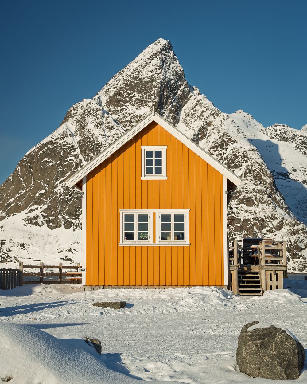 Bright yellow Norwegian cabin standing against a sharp snow-covered mountain peak under clear blue winter sky.