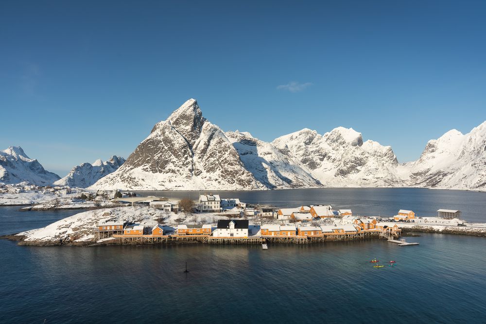 Aerial view of a small fishing village on a rocky peninsula surrounded by snowy mountains in the Lofoten Islands, Norway.