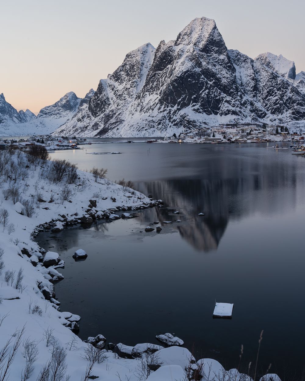 Snow-covered Lofoten Islands fjord with dramatic mountain reflections in calm water during a serene winter twilight in Norway.