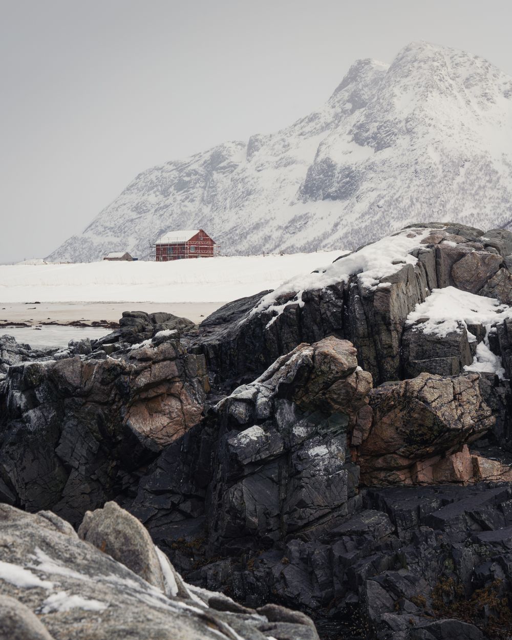 A lone red cabin sits on a snowy plain beneath a massive snow-covered mountain with rugged rocks in the foreground.