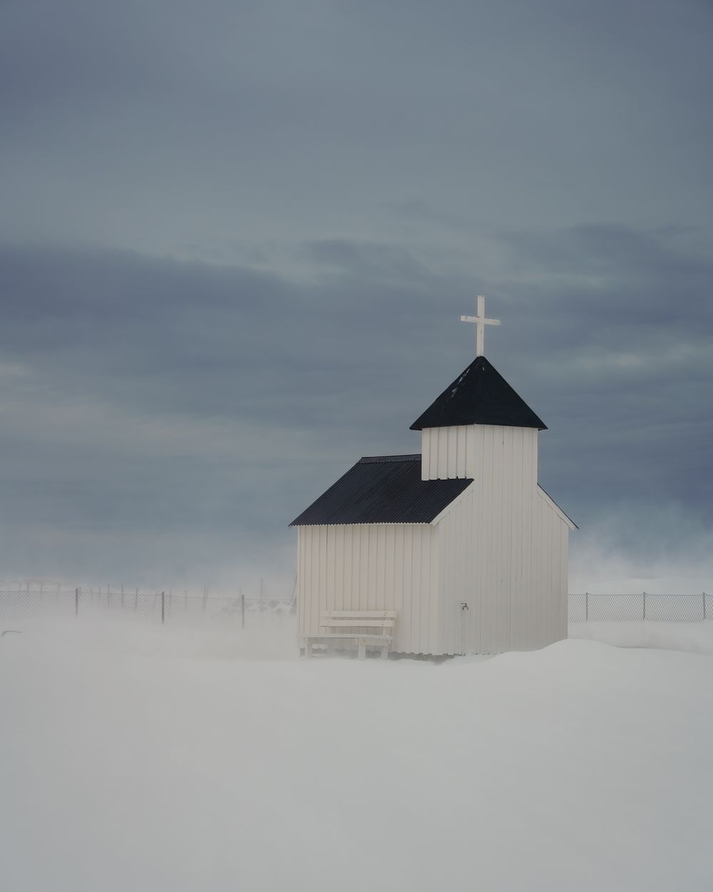 A small white wooden church with a cross stands alone in a vast snowy winter landscape under a cloudy sky.