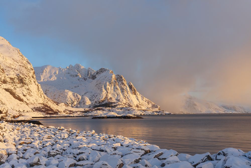 Golden sunrise light illuminating snow-covered rocky mountains and frozen shoreline of a Norwegian fjord in winter.