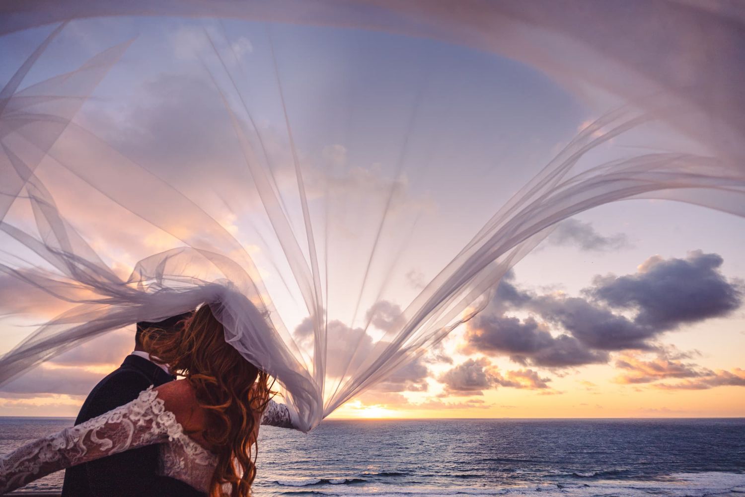 Couple embracing at sunset by the ocean, bride's veil flowing dramatically in the wind against a colorful sky.