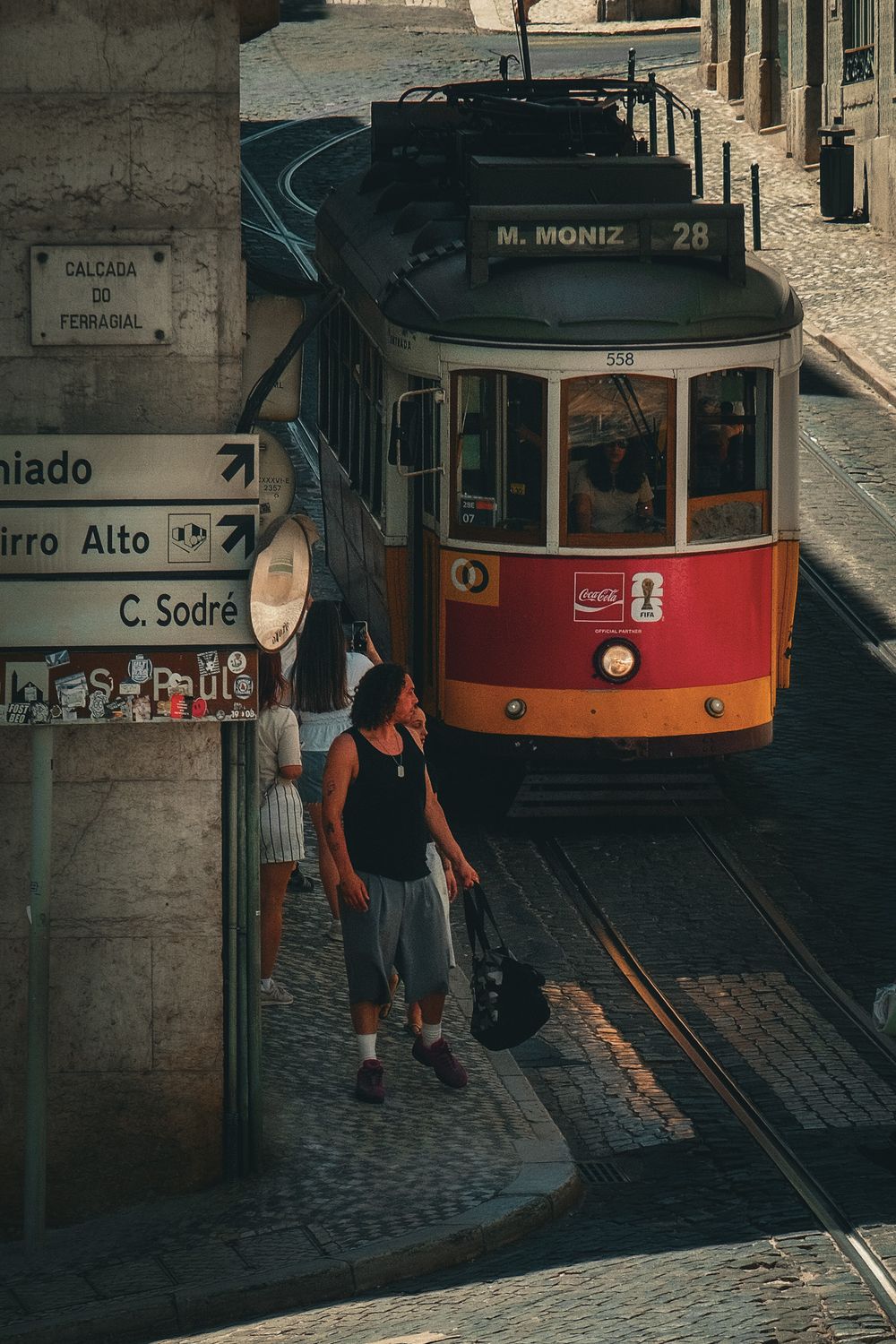 Red tram 28 to M.Moniz on cobblestone street in Lisbon, Portugal, with pedestrians walking alongside historic buildings.