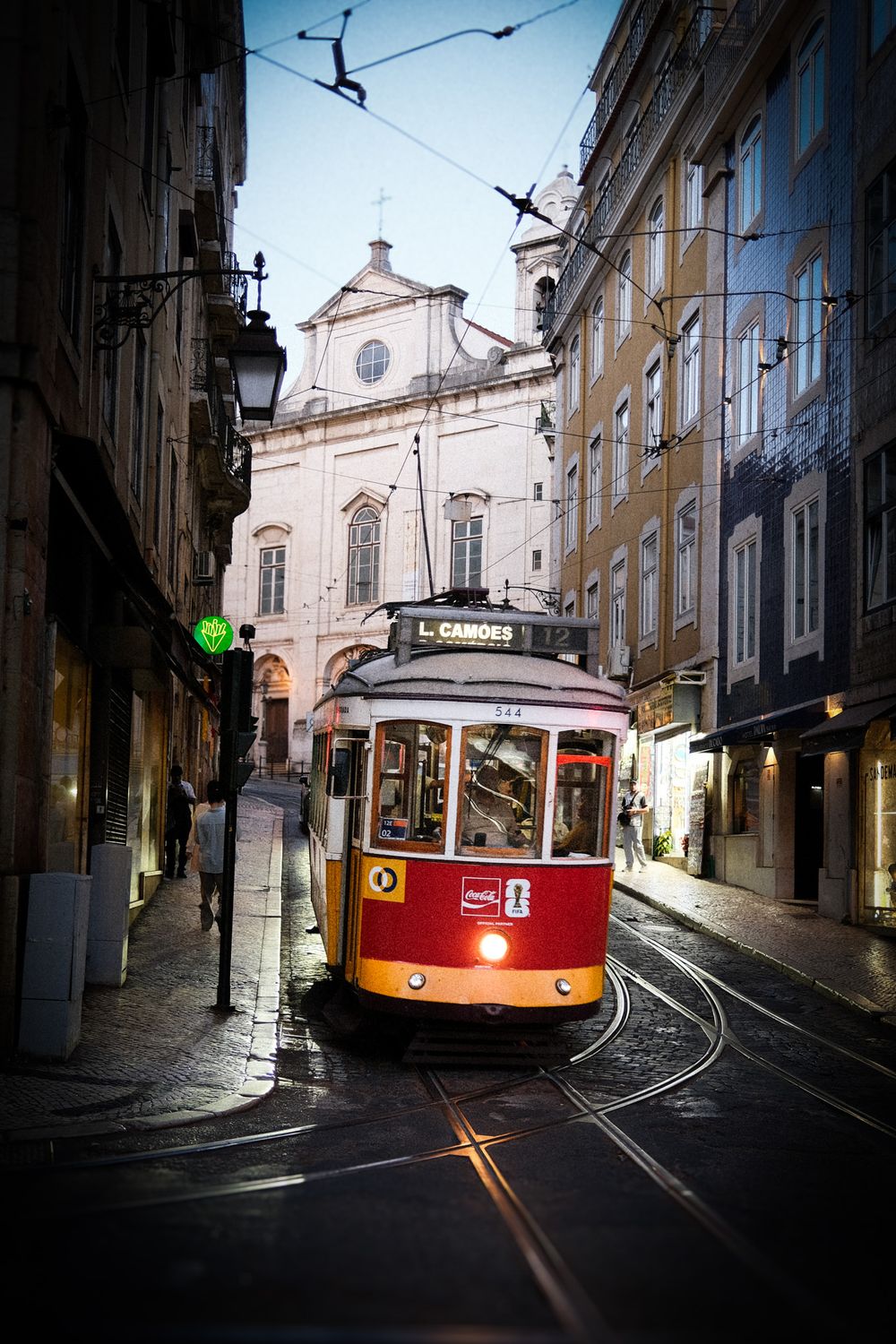 A vintage red and yellow tram navigates a narrow cobblestone street in Lisbon, Portugal, with historic buildings lining the route.