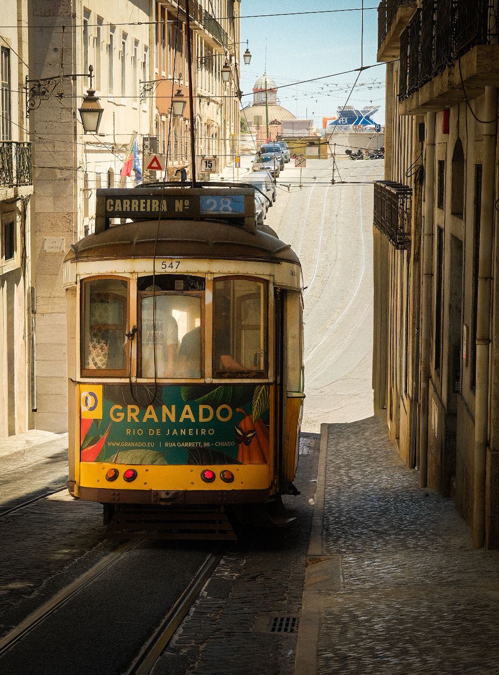 Yellow tram number 28 with Granado Rio de Janeiro advertisement navigating narrow cobblestone streets of Lisbon, Portugal.