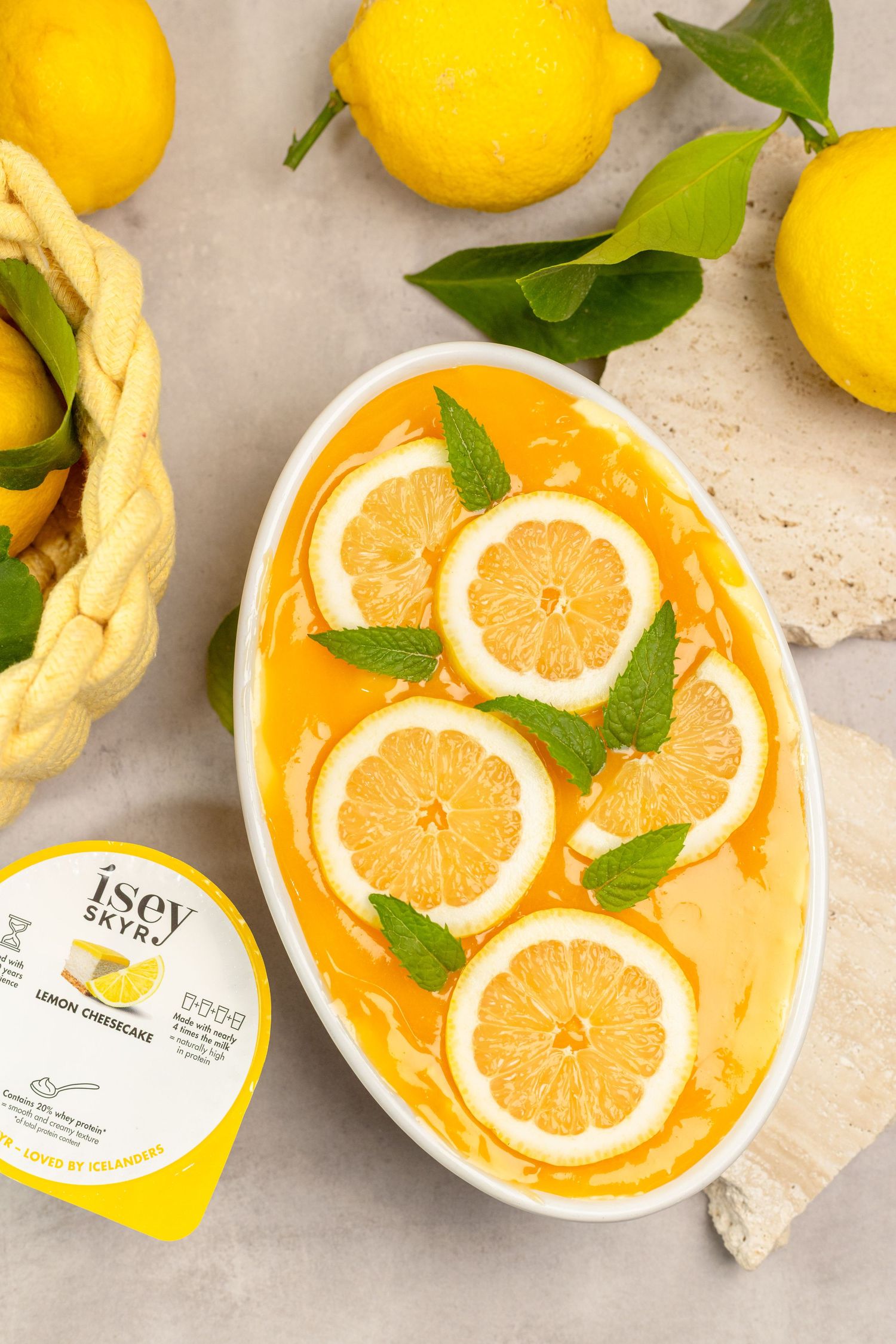 Lemon cheesecake skyr dessert topped with lemon slices and mint leaves, surrounded by fresh lemons on a gray surface.